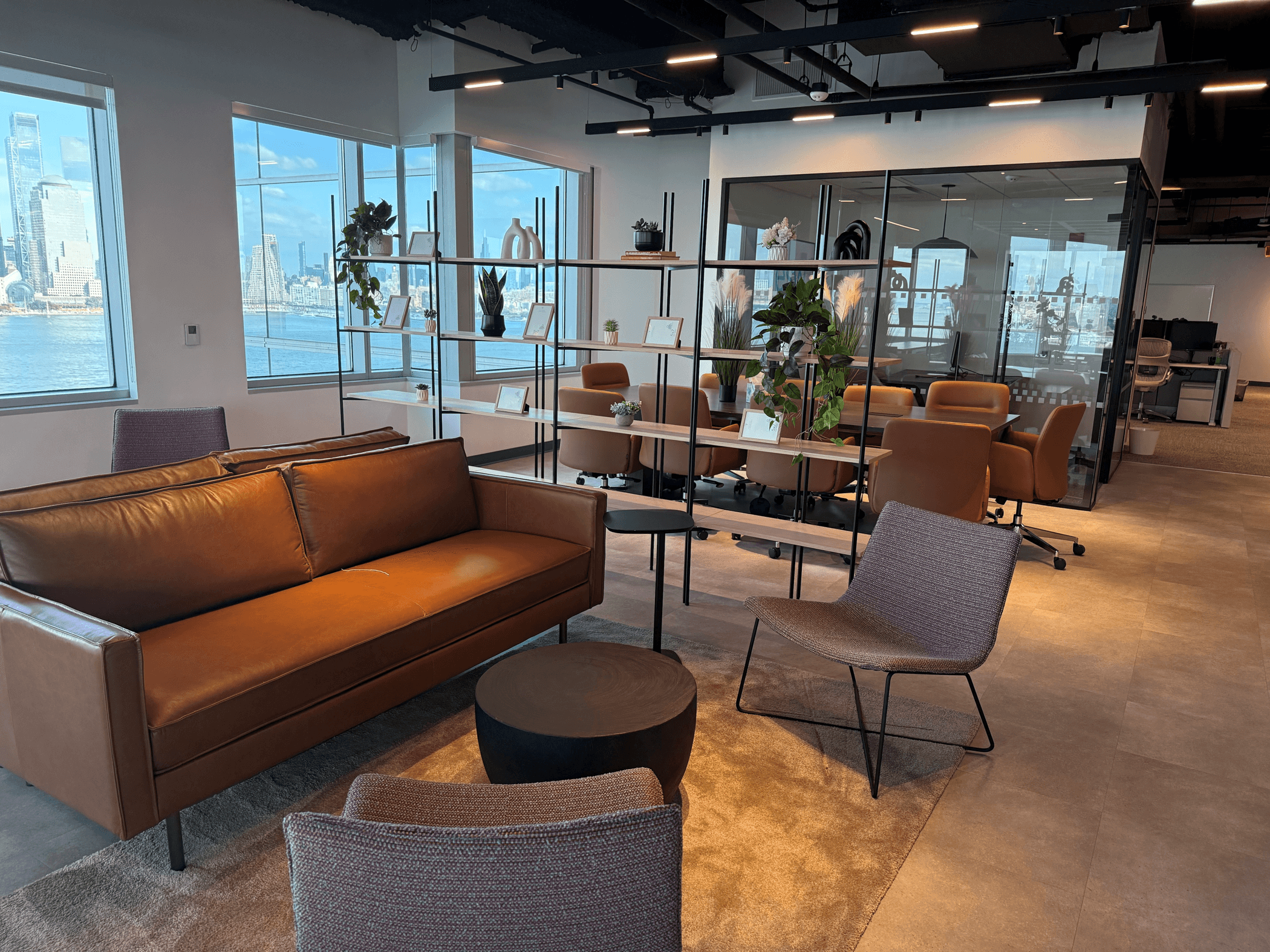 Modern office lounge area with a brown leather sofa, upholstered chairs, and small round tables in the foreground. Behind an open shelving divider with plants and decorative accents, a glass-walled meeting room features tan conference chairs around a large table. Floor-to-ceiling windows overlook a waterfront city skyline, filling the bright, contemporary workspace with natural light.