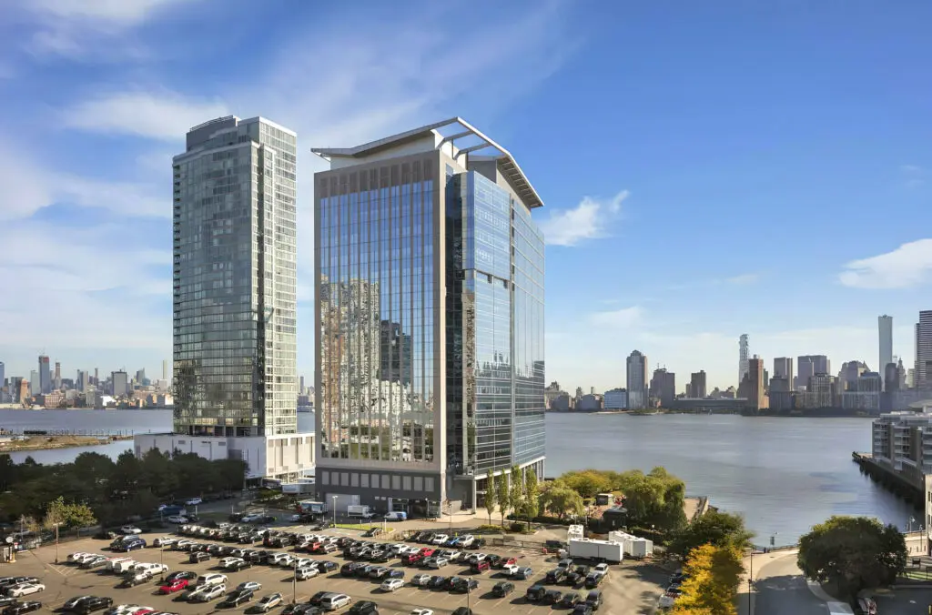 Modern glass office towers situated along a waterfront, with a large parking area in the foreground and a city skyline visible across the river. The reflective high-rise buildings overlook the water under a clear blue sky, surrounded by trees, walkways, and neighboring office and residential developments.