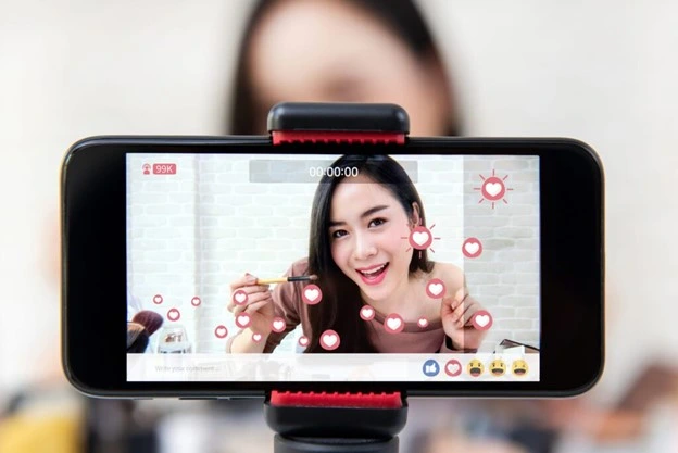 A woman demonstrates makeup application as seen through a cell phone. Hearts and other reactions are on the screen as well.
