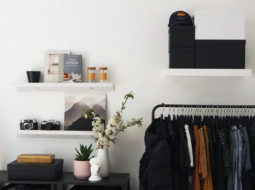 Photo of a minimalist room including clothes on a hanging bar and white shelves with black and white storage boxes. Ecommerce buying trends include clothing and gardening.