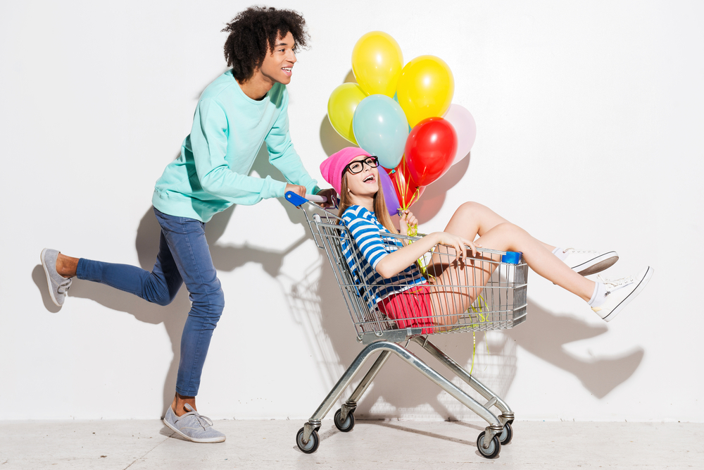 a young man pushes a young woman holding balloons in a shopping cart